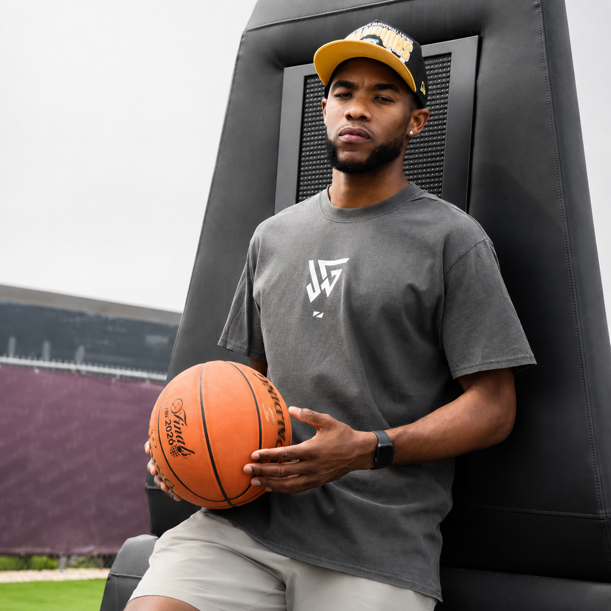 A man wearing a dark gray t-shirt with a white graphic logo, beige shorts, and a yellow and black cap sits on a black padded training chair holding an orange basketball.
