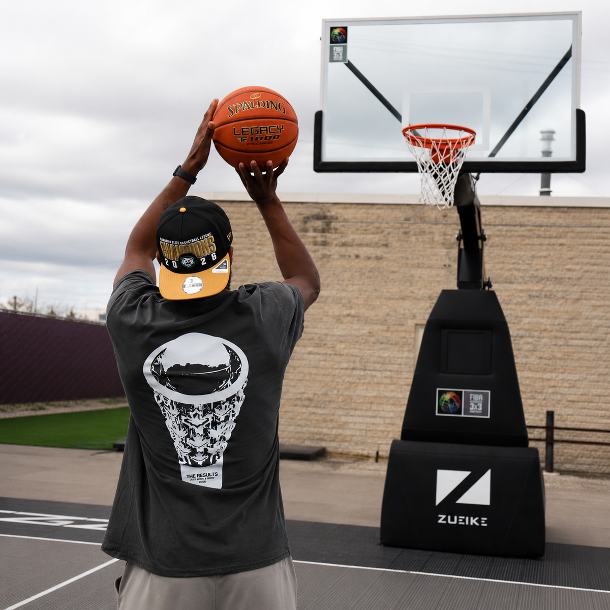 A man wearing a black and orange baseball cap and a black graphic t-shirt holds an orange Spalding basketball above a Zueik portable basketball court setup with a clear backboard.