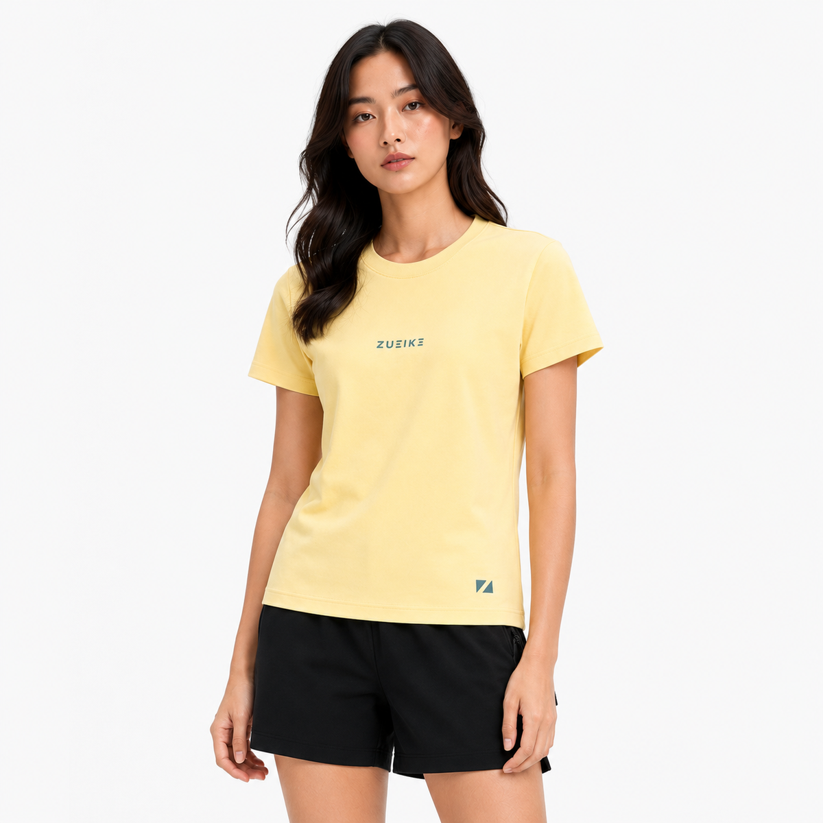 A woman modeling a pale yellow short-sleeve athletic T-shirt with 'ZUEIKE' printed in green on the chest and a small green logo near the lower hem, paired with black shorts against a light gray studio background.