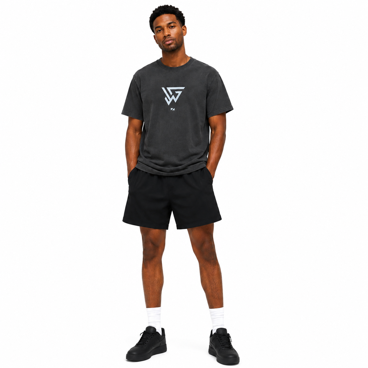A man modeling a dark gray washed t-shirt with a white geometric logo on the chest, paired with black athletic shorts, white socks, and black sneakers, set against a plain white background.